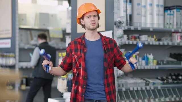 Cheerful millennial in hard hat imitating shooting at camera with caulking gun. Joyful young Caucasian man having fun buying tools in hardware store. Lifestyle and industry concept.