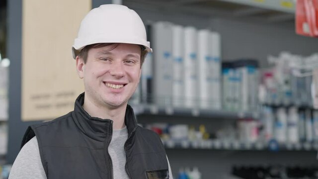Close-up portrait of young positive Caucasian man in hard hat posing in hardware store with blurred shelves at background. Happy worker looking at camera smiling. Service and occupation.