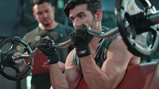Caucasian man using barbell for weight training with trainer in gym.