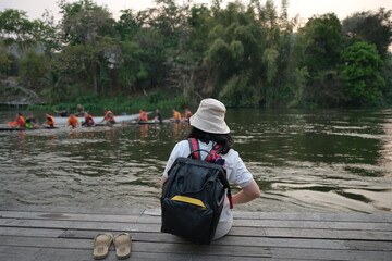 Asian woman in a white T-shirt, black backpack, wearing hat and taking off shoes, sits comfortably on a wooden bridge or pier to watch tourists ride a boat or Swimming in the river in evening sunset