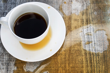 A white cup sits on a saucer in a puddle of spilled coffee on a wooden table.