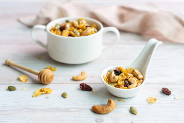 Close up of homemade honey caramel cornflakes in white bowl with cashew nut, pumpkin seeds and dried raisins on white wooden background. Grains cornflakes for healthy food concept. Flat lay