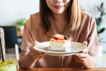 Smiling Asian girl holding a plate of her favorite strawberry cheese cake on wooden table in cafe. Food and dessert concept.