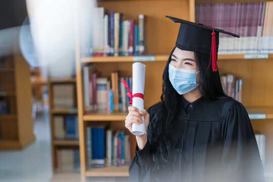 A young cheerful Asian woman university graduate in graduation gown and cap wears a sanitary mask holds a degree certificate to celebrate her education achievement on the commencement day. Stock photo