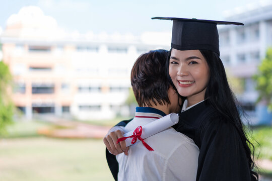 Young happy excited Asian woman university graduates in graduation gown and cap with a degree certificate hugs a friend to celebrate her education achievement on the commencement day. Stock photo