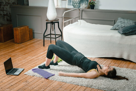 A young woman goes in for sports at home, online workout . The athlete  pumps up the buttocks, makes the gluteal bridge in the bedroom, in the background there is a bed, a vase, a carpet.