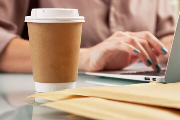 Young woman drinking coffee from disposable cup at the office while working with a computer. Brown paper cup with a white plastic lid 