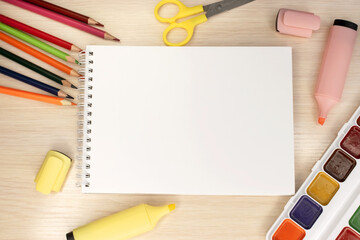 On the wooden table among the school supplies is a notebook with a blank white sheet. Paint and colored pencils with scissors and marker. Template and banner. Training
