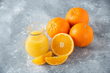 A glass pitcher of juice with fresh orange fruits on stone background