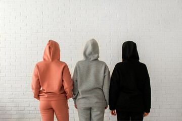 Three girls in warm fleece tracksuits stand with their backs to a white brick wall. Sports...