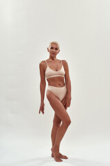 Full length shot of attractive caucasian mature woman in underwear looking at camera while posing half naked in studio against light background