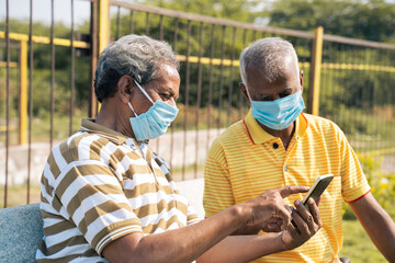 Side view Selective focus on mobile, elderly man in medical face mask showing mobile phone to his old friend at park - concept of senior people using smartphone, technology, internet and social media