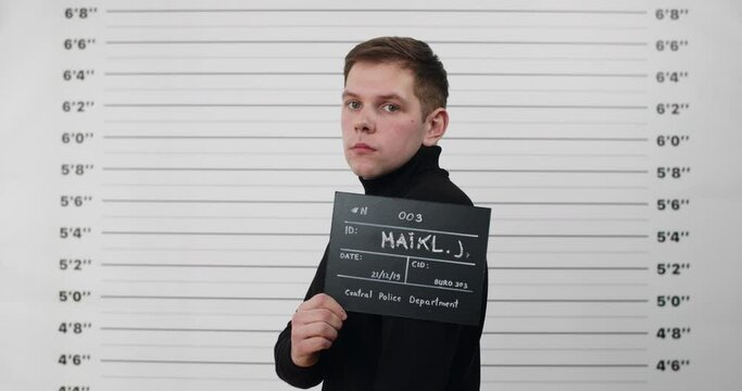 Mugshot of caucasian guy turning head and looking to camera while standing aside . Crop view of criminal man in black turtlrneck holding sign and posing for photo in police department.