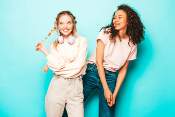 Two young beautiful smiling international hipster female in trendy summer clothes. Sexy carefree women posing near blue wall in studio. Positive models having fun. Concept of friendship