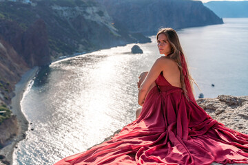 A girl with loose hair in a red dress sits on a rock rock above the sea. In the background, the sun...