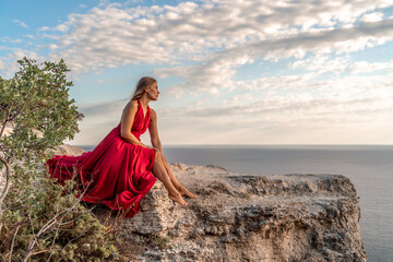 A girl with loose hair in a red dress sits on a rock rock above the sea. In the background, the sea. The concept of travel.