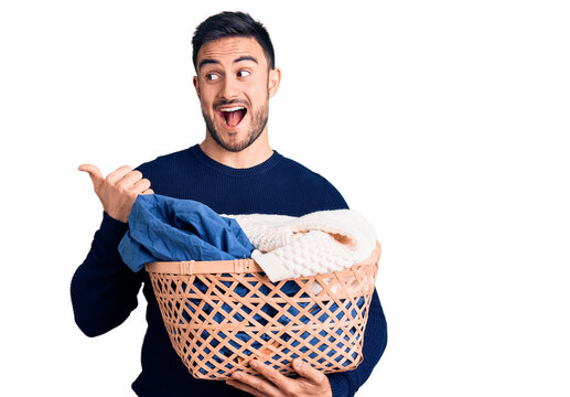 Young handsome man holding laundry basket pointing thumb up to the side smiling happy with open mouth