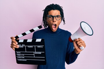 Young african american man holding video film clapboard and megaphone in shock face, looking...