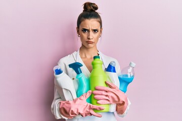Beautiful brunette young woman wearing cleaner apron holding cleaning products skeptic and nervous,...