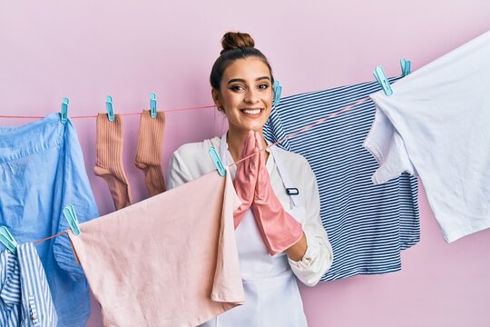 Beautiful brunette young woman washing clothes at clothesline praying with hands together asking for forgiveness smiling confident.