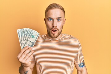 Young caucasian man holding dollars scared and amazed with open mouth for surprise, disbelief face