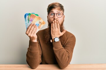 Young irish redhead man holding swiss franc banknotes sitting on the table covering mouth with...