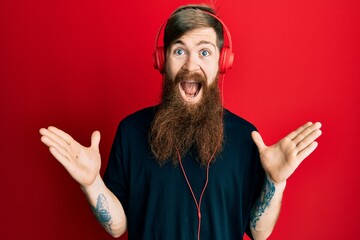 Redhead man with long beard listening to music using headphones celebrating crazy and amazed for success with arms raised and open eyes screaming excited. winner concept