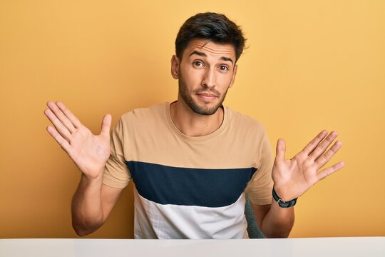 Young handsome man wearing casual clothes sitting on the table clueless and confused with open arms, no idea and doubtful face.