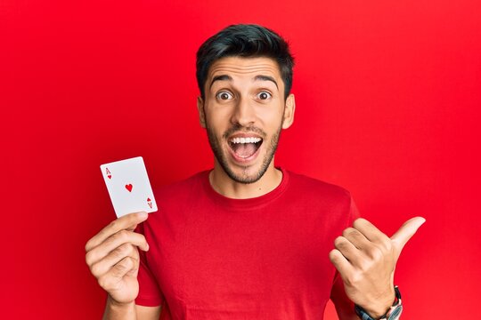 Young handsome man holding ace poker card pointing thumb up to the side smiling happy with open mouth