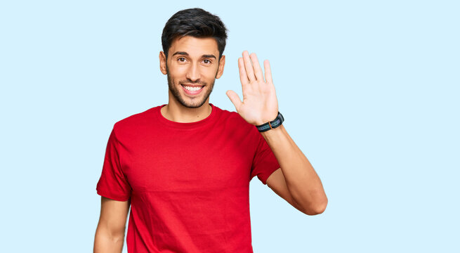 Young handsome man wearing casual red tshirt waiving saying hello happy and smiling, friendly welcome gesture