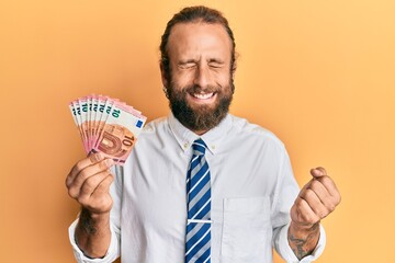 Handsome business man with beard and long hair holding bunch of 10 euro banknotes screaming proud, celebrating victory and success very excited with raised arm