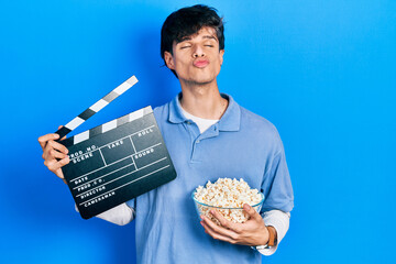 Handsome hipster young man eating popcorn holding cinema clapboard looking at the camera blowing a...