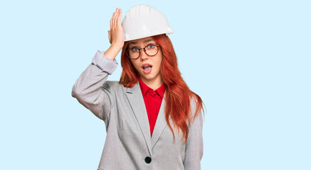 Young redhead woman wearing architect hardhat surprised with hand on head for mistake, remember error. forgot, bad memory concept.