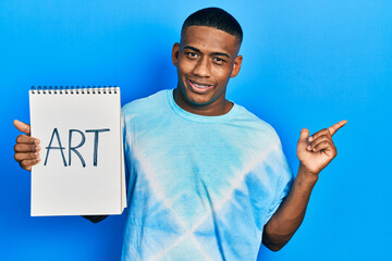 Young black man holding art notebook smiling happy pointing with hand and finger to the side