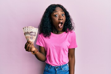 Beautiful african young woman holding 500 mexican pesos banknotes scared and amazed with open mouth for surprise, disbelief face