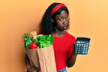 Beautiful african young woman holding groceries and calculator in shock face, looking skeptical and...