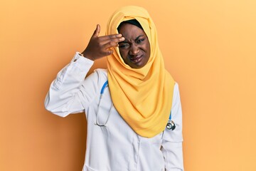 Beautiful african young woman wearing doctor uniform and hijab pointing unhappy to pimple on forehead, ugly infection of blackhead. acne and skin problem