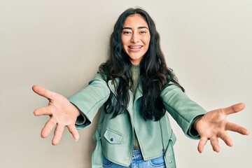 Hispanic teenager girl with dental braces wearing green leather jacket looking at the camera smiling with open arms for hug. cheerful expression embracing happiness. - obrazy, fototapety, plakaty