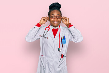 Young african american woman wearing doctor uniform and stethoscope smiling pulling ears with...