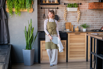 Happy woman in a linen apron standing in kitchen by sink wiping a plate with a linen towel