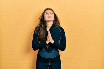 Young hispanic girl wearing diver neoprene uniform begging and praying with hands together with...