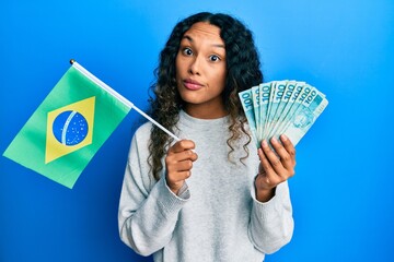 Young latin woman holding brazil flag and real banknotes in shock face, looking skeptical and...