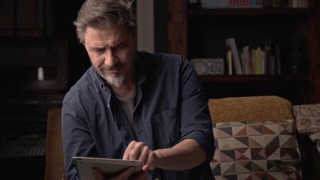 Man sitting on couch at home with tablet computer. Businessman working in home office. Portrait of mature age, middle age, mid adult man, bearded, authentic look.