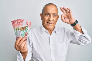 Handsome senior man holding 100 new zealand dollars banknote doing ok sign with fingers, smiling...
