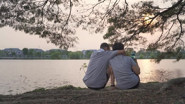 happy senior couple sitting near lake view hugging at tree in evening sunset. smiley sweet asian elderly lover outdoor romantic sharing good moment together. sport mature in sportswear workout at park