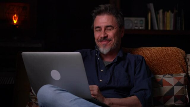 Man sitting on couch at home with laptop computer, talking on video chat. Businessman working in home office. Portrait of mature age, middle age, mid adult man, bearded, authentic look.