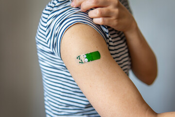 Woman showing her arm with a fun, colorful, adhesive bandage after injection of vaccine or a scratch on the skin. First aid. Medical, pharmacy, and healthcare concept. After vaccination treatment.
