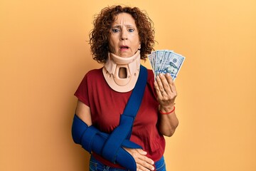 Beautiful middle age mature woman wearing cervical collar and sling holding insurance dollars in...
