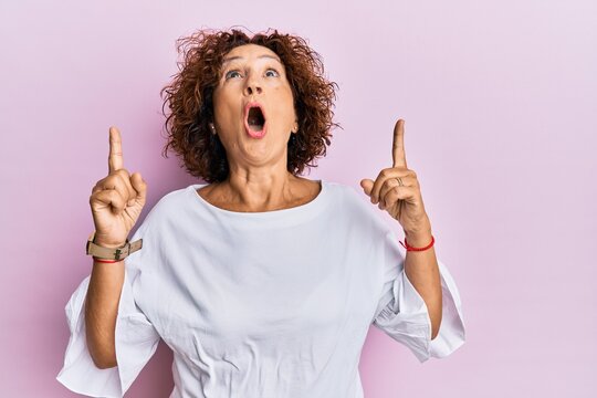 Beautiful middle age mature woman wearing elegant clothes over pink background amazed and surprised looking up and pointing with fingers and raised arms.