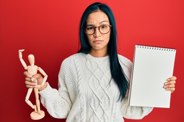 Beautiful hispanic woman holding small wooden manikin and notebook skeptic and nervous, frowning...